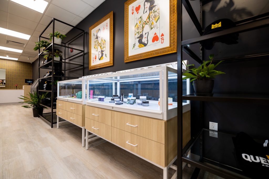 A display of cannabis products inside Queen City Dispensary in Plainfield, NJ. A showcase in the center with a glass top and a light brown bottom contains cannabis flower and preroll packaging. Art inspired by the Queen of Diamonds and the Queen of Hearts hangs above the display on a dark wall.