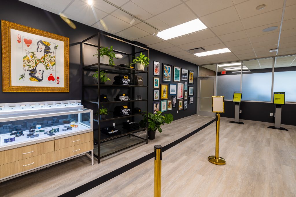 Queen City Dispensary interior showing a large framed playing card, accessories display, black shelf with merchandise, framed art wall, kiosks and gold stanchions.