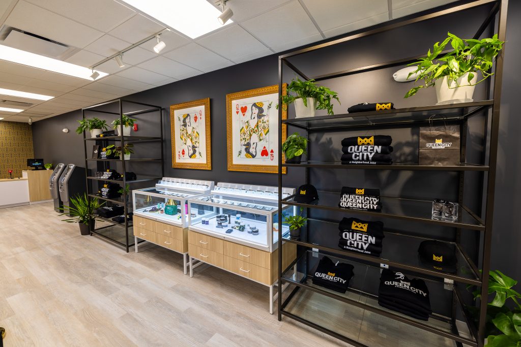 A display of cannabis products inside Queen City Dispensary in Plainfield, NJ. A showcase in the center with a glass top and a light brown bottom contains cannabis flower and preroll packaging. Art inspired by the Queen of Diamonds and the Queen of Hearts hangs above the display on a dark wall.