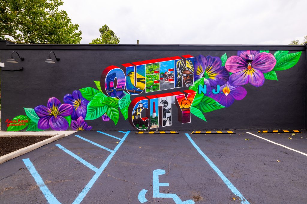 The Queen City mural painted by artist Danielle Mastrion appears on a black background. Queen City is painted in vintage lettering, and each letter features a different attribute connected to Plainfield, Union County, and New Jersey. The mural is surrounded by vibrant green leaves and purple violets.