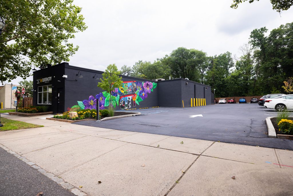An image of Queen City Dispensary in Plainfield, NJ from the outside. The photo is taken from South Avenue from the angle of the parking lot. The Queen City mural painted by Danielle Mastriano can be seen in the photo, painted onto a black one-story building. A small tree can be seen in front of the mural.