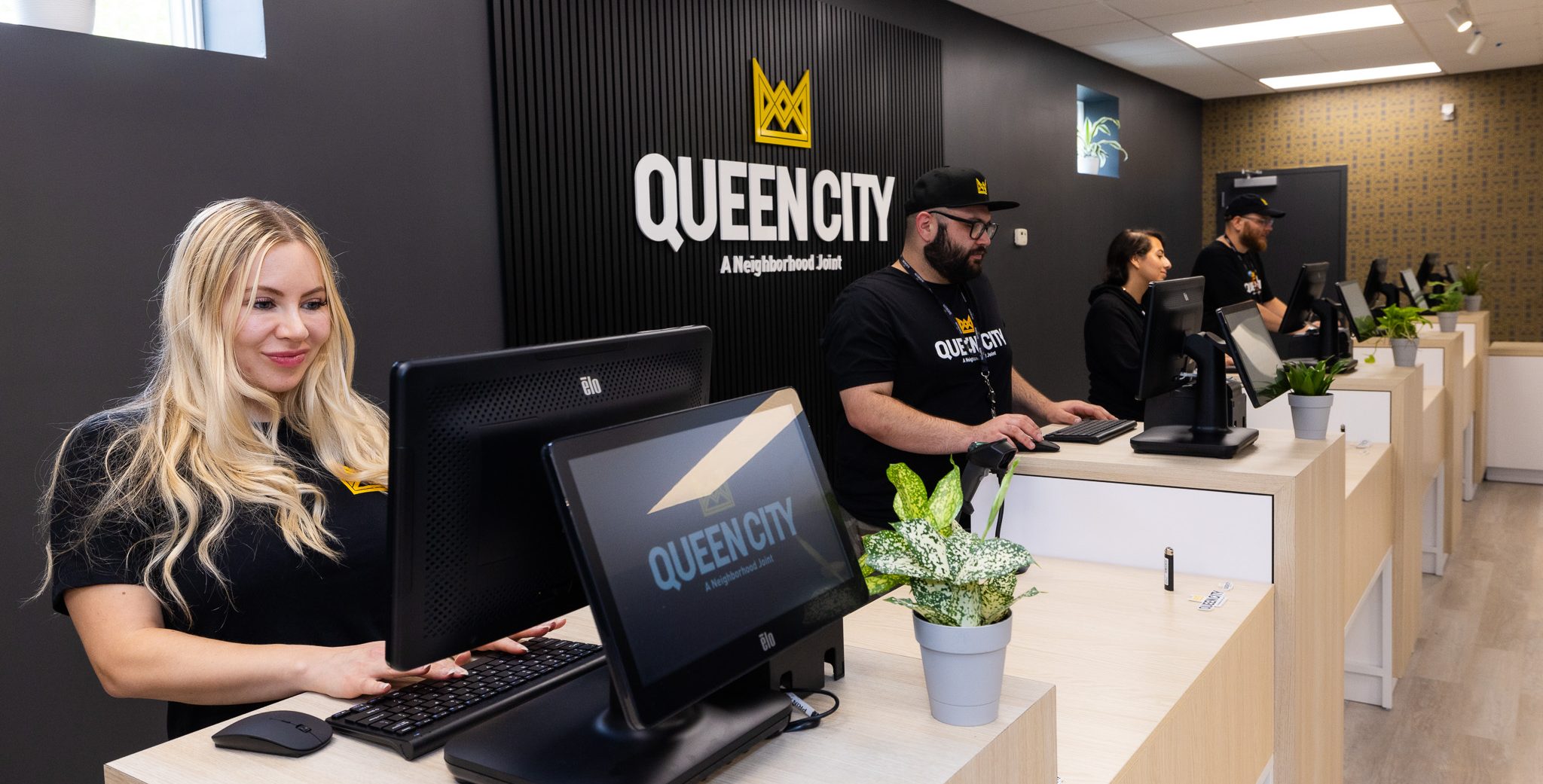 Four budtenders are standing at the checkout counter at Queen City in Plainfield, New Jersey, each behind a checkout station. Two of the budtenders are sporting beards and hats. All four budtenders are wearing black and branded Queen City t-shirts. The store's logo is behind them.