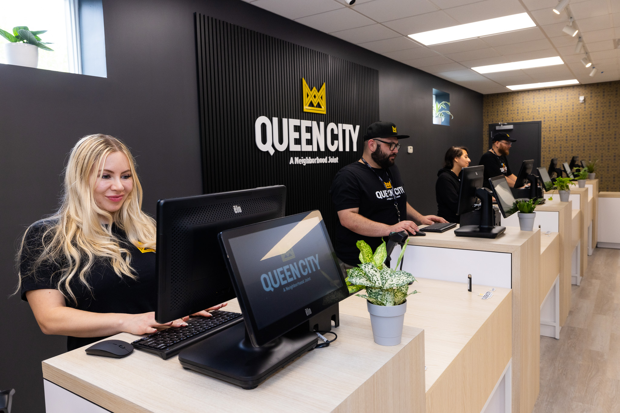 Four budtenders are standing at the checkout counter at Queen City in Plainfield, New Jersey, each behind a checkout station. Two of the budtenders are sporting beards and hats. All four budtenders are wearing black and branded Queen City t-shirts. The store's logo is behind them.