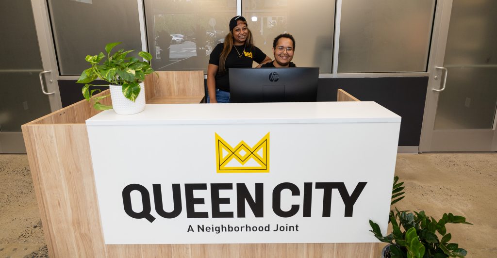 Two Queen City Dispensary staff members smiling and sitting at a tan desk behind a white sign with black letters displaying the Queen City logo.