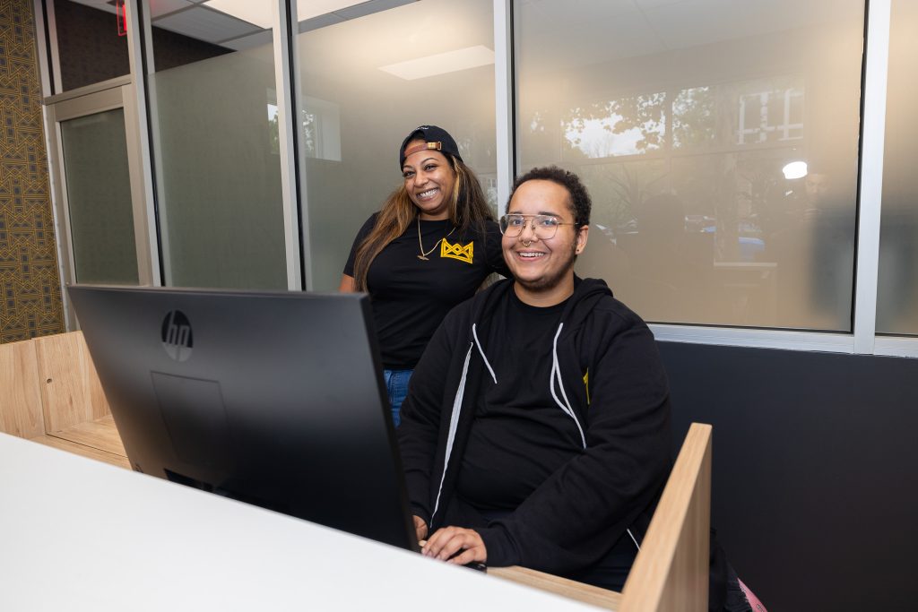 Two Queen City Dispensary staff members smiling and sitting at a tan desk behind an HP computer monitor.