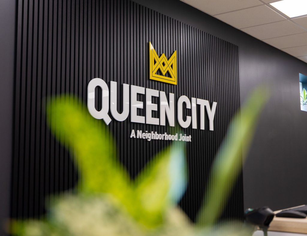 Queen City Dispensary sign on black wall with blurred leaves in the foreground