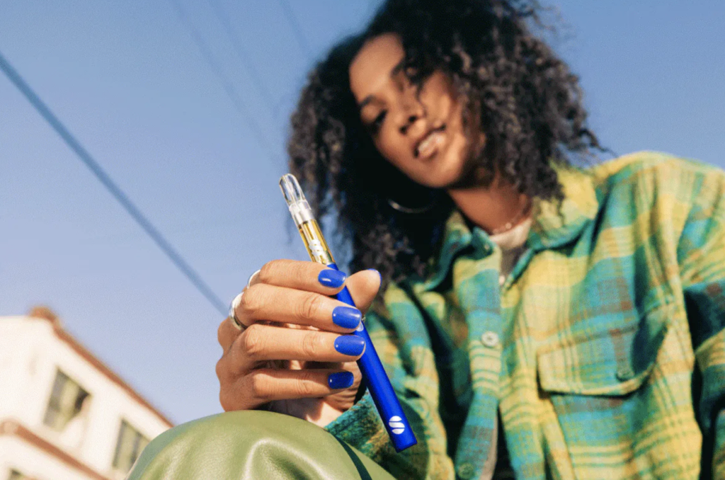 Person with black hair, green and teal plaid shirt, and blue nails holding blue cannabis vape pen