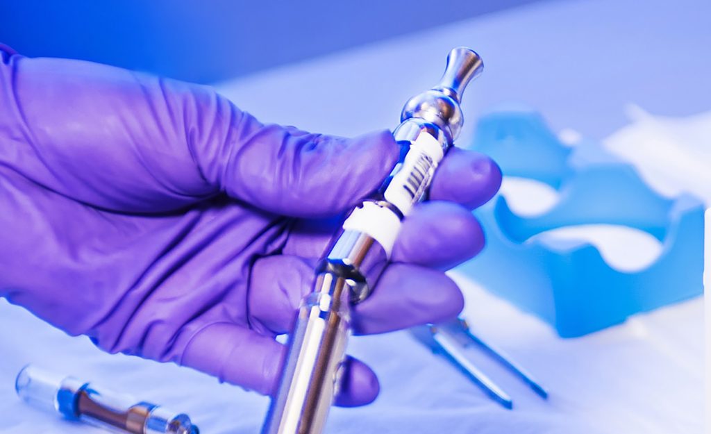 Hand with purple latex glove holding a silver cannabis vape cartridge and battery in front of blue background