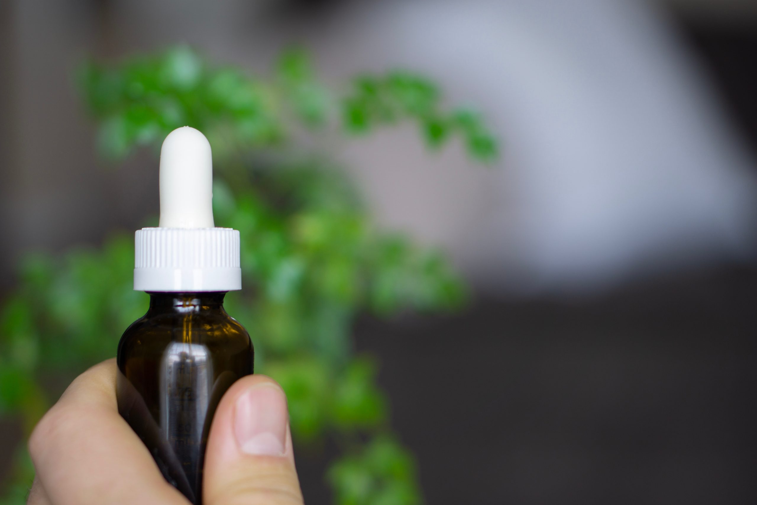 A close up of a hand holding a brown tincture bottle with a white top. Green plants are blurred in the background.