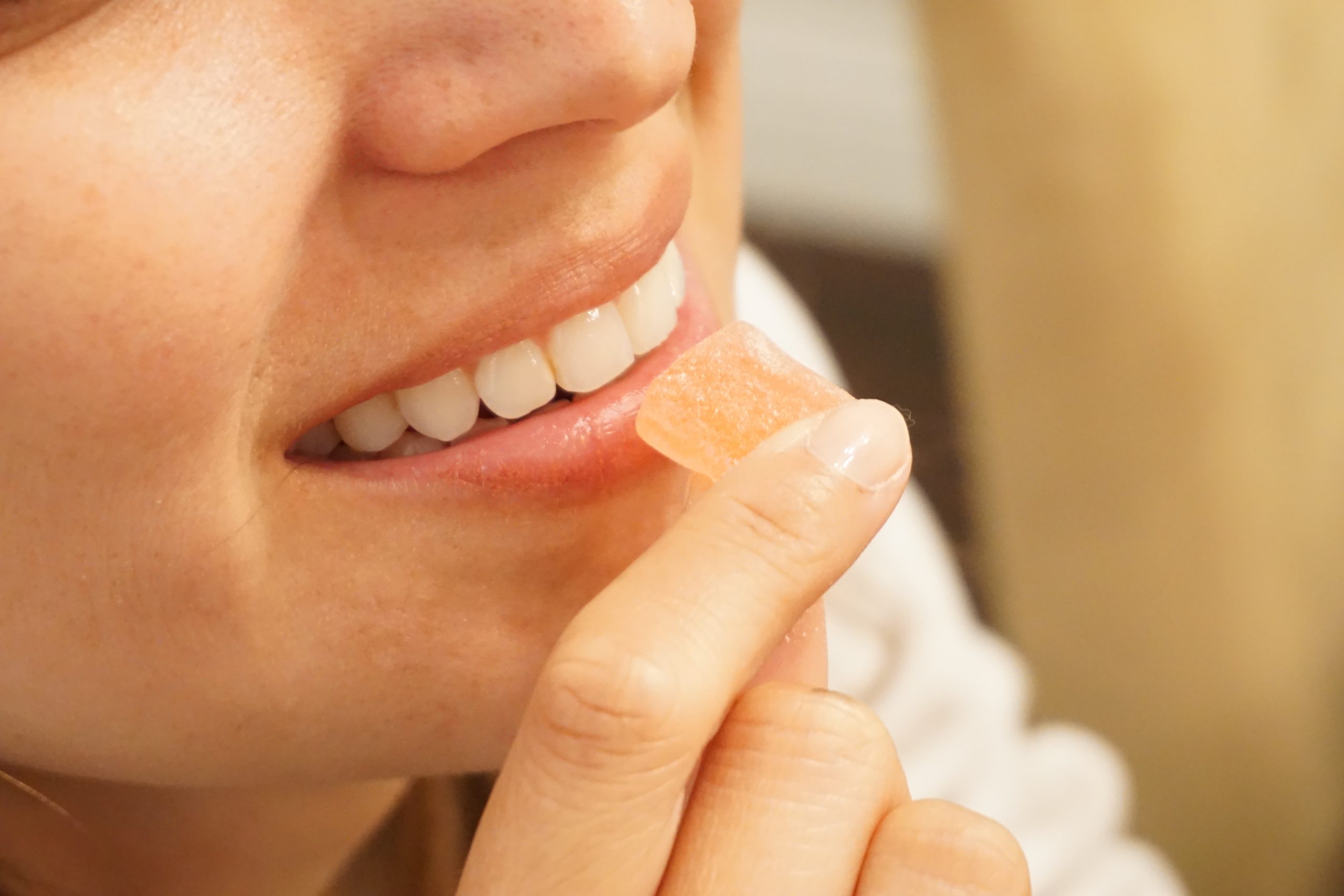 A close up photo of a smiling person holding a square gummy. The gummy is held up to her lips.