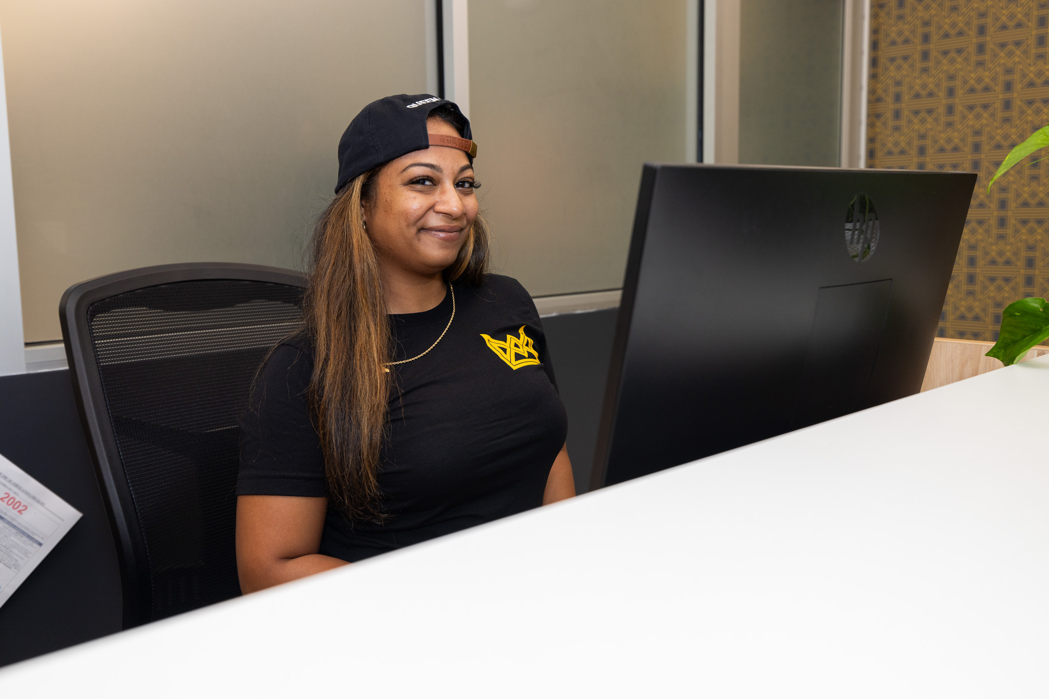 Queen City Dispensary Assistant General Manager Ariell Hunt poses behind the check-in desk at Queen City. She is sitting in a chair and wearing a black baseball cap backward. She is wearing a black t-shirt with the Queen City logo on it. Her hair is straight.