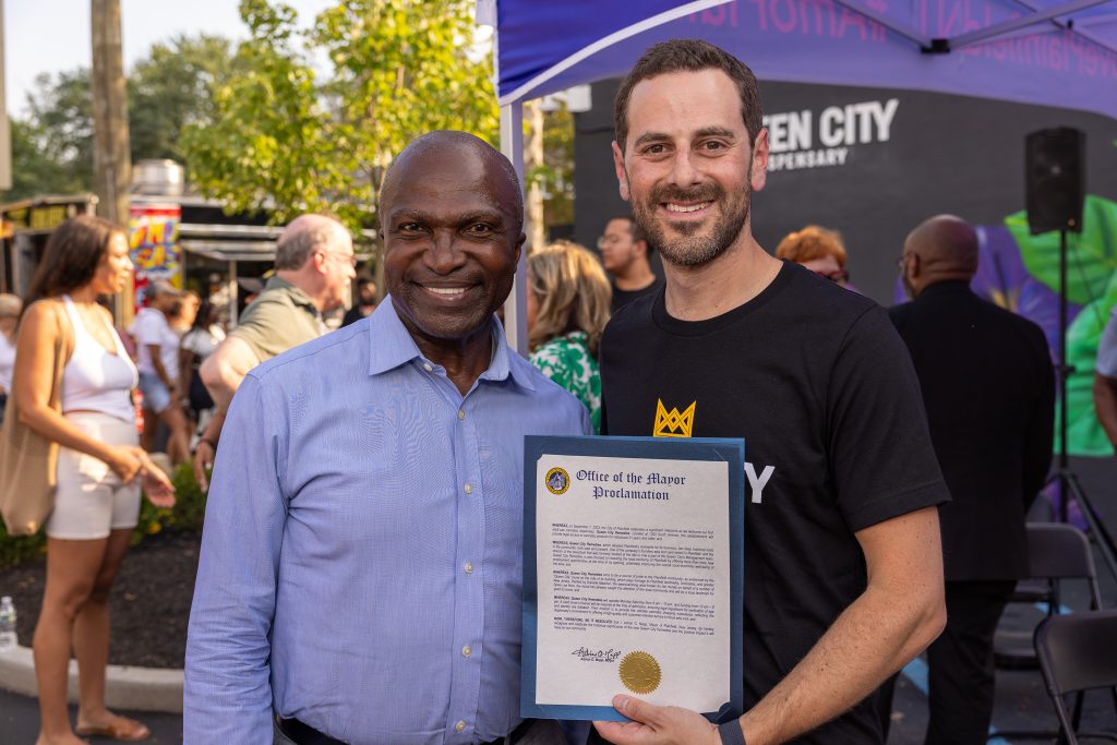 Plainfield NJ Mayor Adrian O Mapp is on the left, wearing a purple shirt. He is standing with with Queen City Dispensary team member Justin Singer. Justin is wearing a Queen City t-shirt. He is holding a proclamation from the city of Plainfield declaring September 7th Queen City Remedies Day in Plainfield.