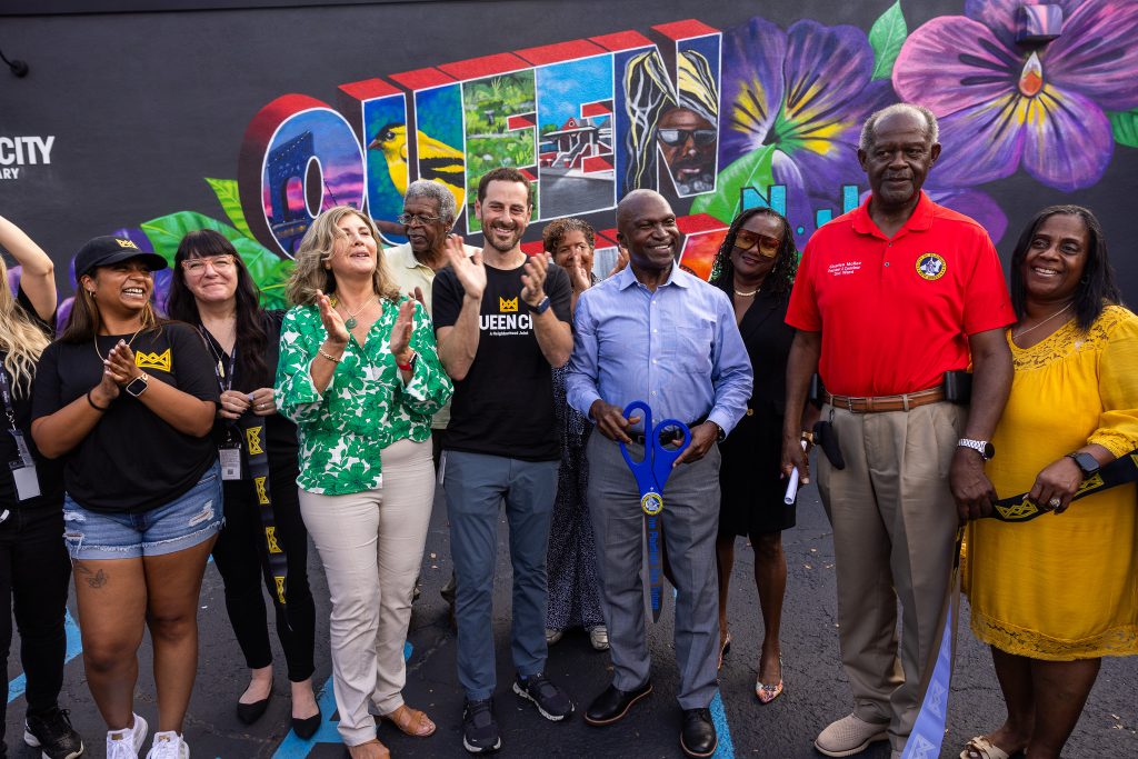 Queen City team members and officials from the city of Plainfield pose after the official ribbon cutting. Queen City team members are standing on the left, while Plainfield city officials are standing on the right. They pose in front of a colorful mural that says "Queen City."