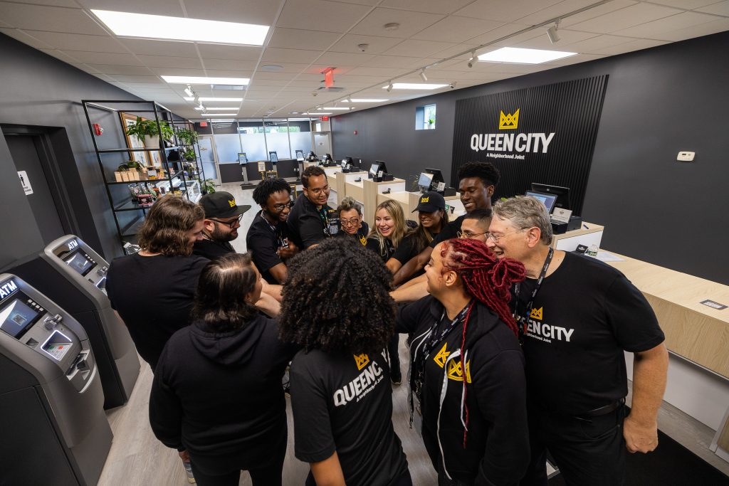 Queen City team members stand inside the dispensary before opening. They are standing in a circle with their hands in the middle. The team members represent different races and ages. All team members are wearing Queen City t-shirts.