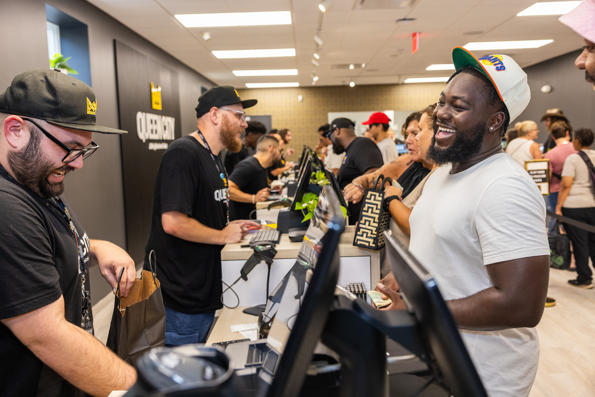 A customer and budtender converse at checkout. The customer on the right is wearing a white T-shirt and hat. The budtender on the left is wearing Queen City gear. Everyone is smiling and laughing, indicating a great experience at a New Jersey recreational weed shop.
