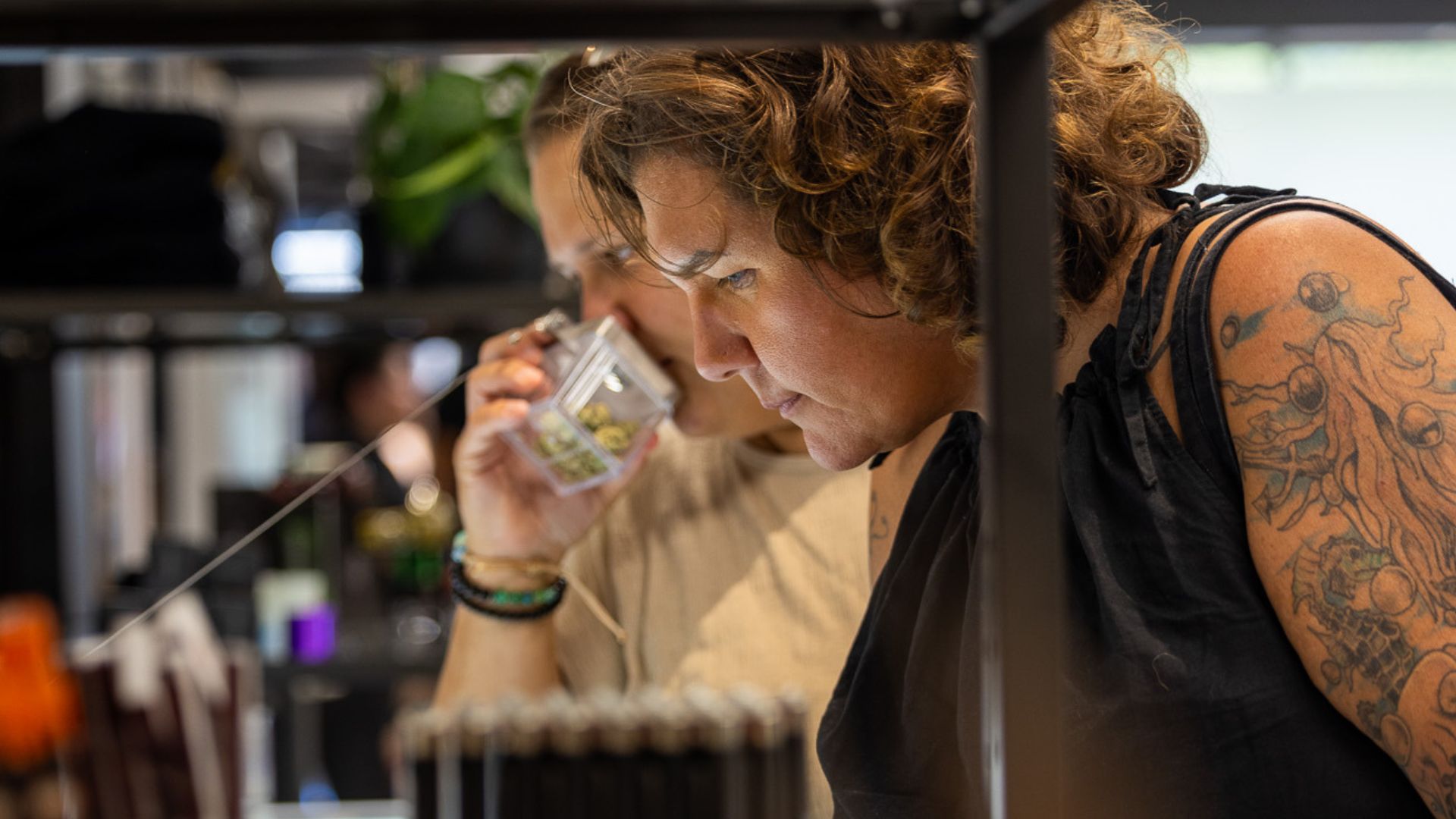 Two Queen City customers examine cannabis flower at the display. The person is the foreground is wearing a black shirt. The person is the background is wearing a tan shirt and is smelling a jar of cannabis.