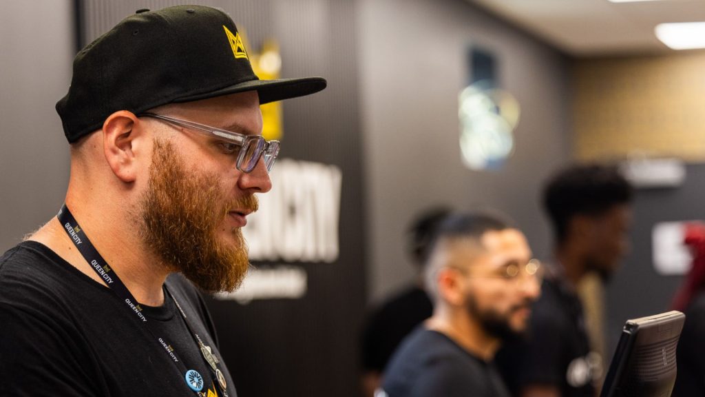 A Queen City associate stands behind the counter while assisting a customer. The budtender has a beard and is wearing a black hat, a black Queen City t-shirt, and an employee ID. His hand is raised as he uses the computer to complete the checkout process for the customer.