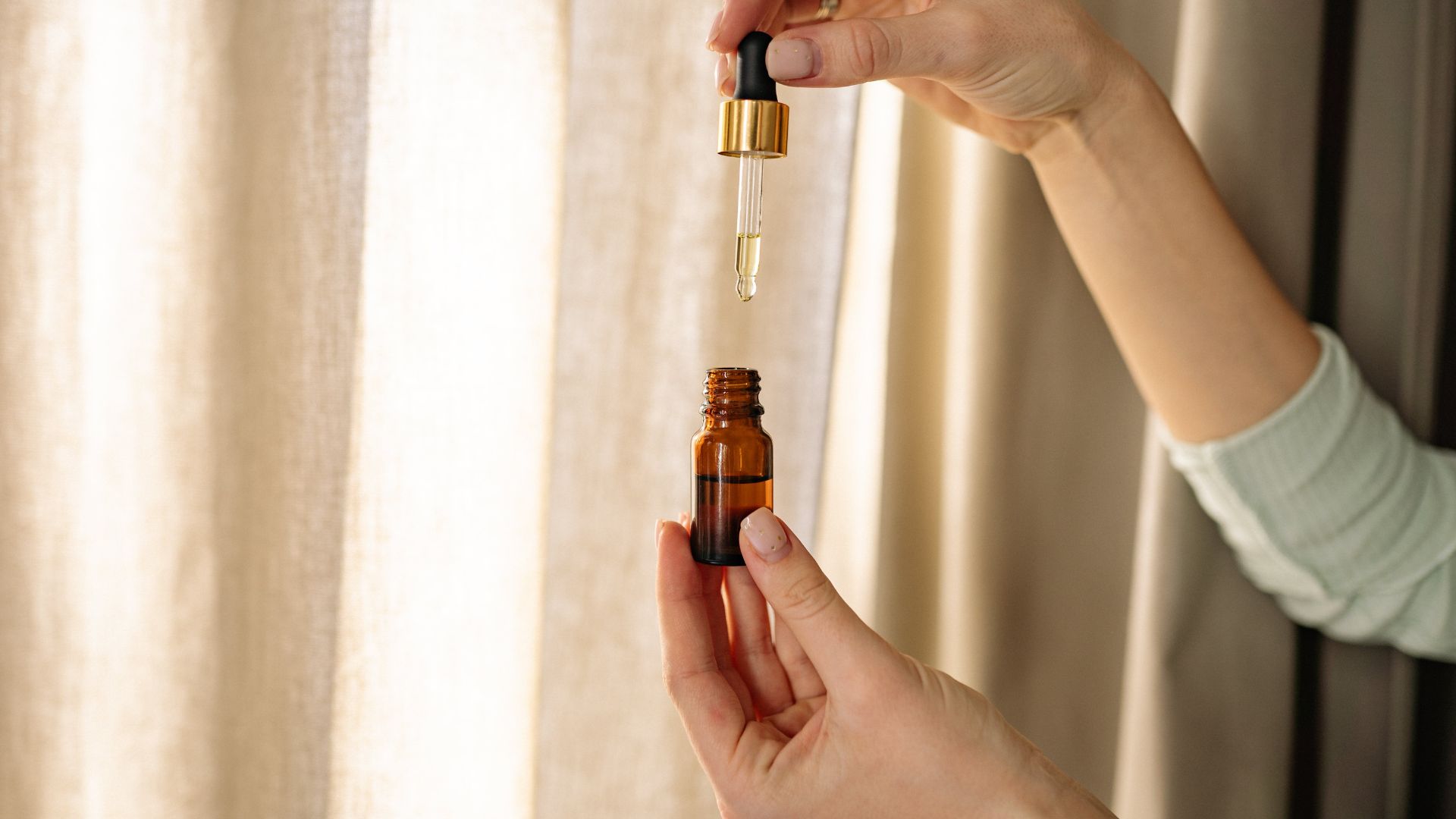 A person holds a tincture bottle and dropper in front of a window with drapes.