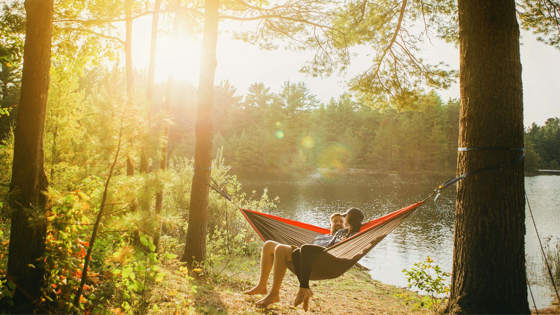 Two people sit on a hammock hanging between two trees alongside a body of water. They are quite relaxed, perhaps after enjoying some cannabis.