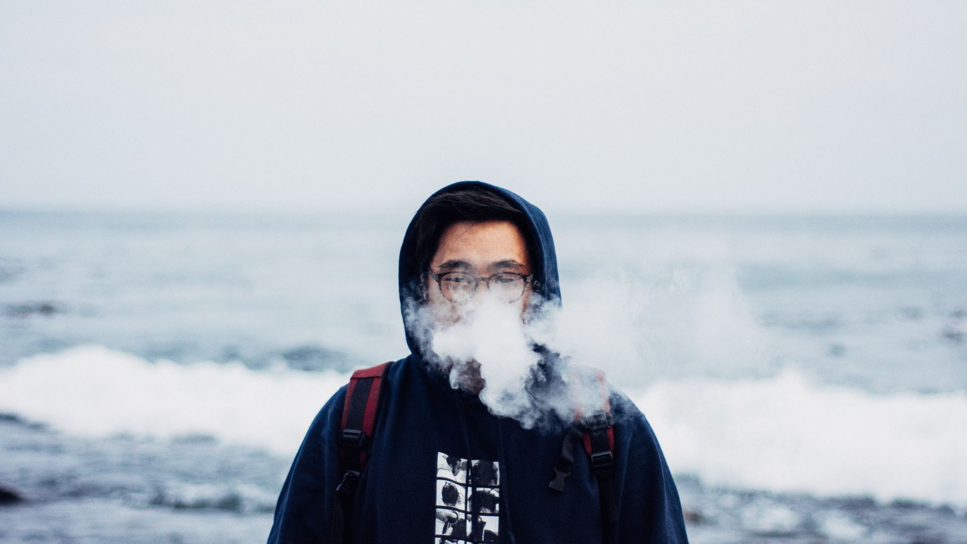 Person in a black hoodie and glasses vaping against an ocean backdrop. Perhaps this person just picked up one of the best vape carts in New Jersey after a trip to Queen City!