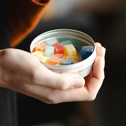 Hand holding round tin filled with red, yellow, orange, blue, and green cubed edibles.