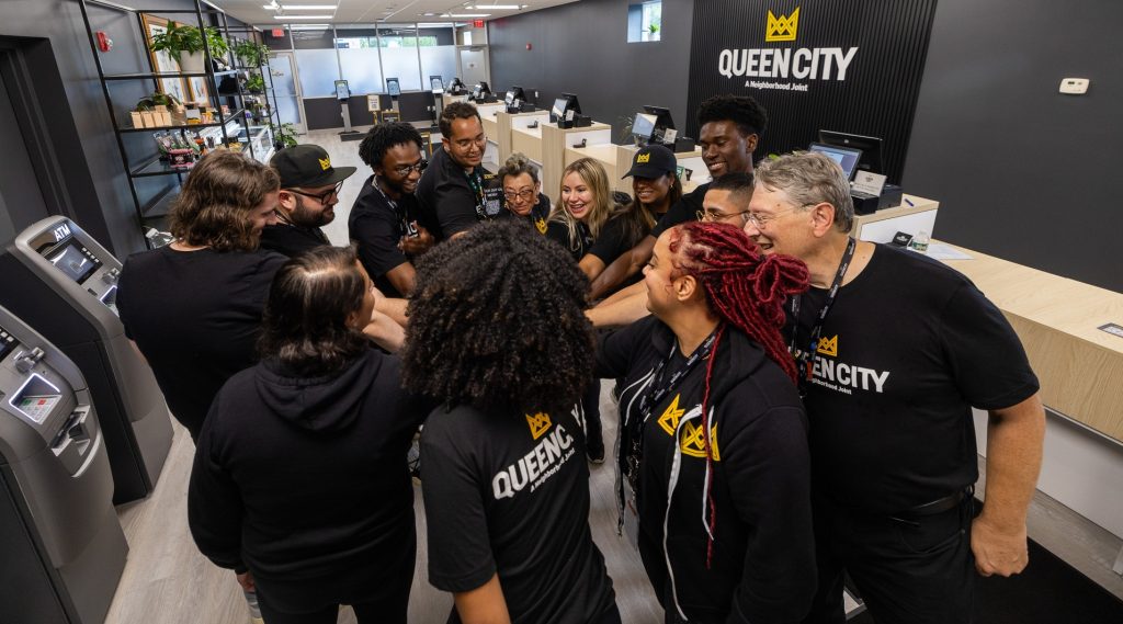 Queen City team members stand inside the dispensary before opening. They are standing in a circle with their hands in the middle. The team members represent different races and ages. All team members are wearing Queen City t-shirts.