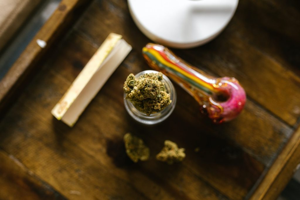 Top view of cannabis in a jar sitting next to a colorful pipe and rolling papers on wooden surface