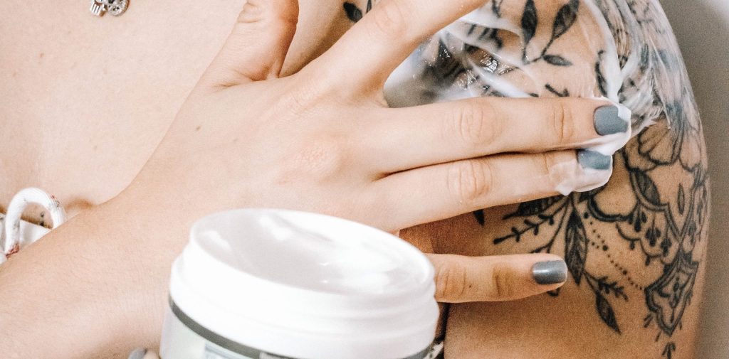 Person with gray nail polish applying white cannabis topical to tattooed shoulder