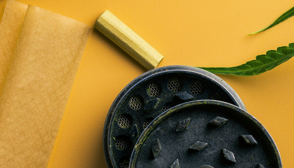 Rolling paper, black grinder, and cannabis leaf on yellow background