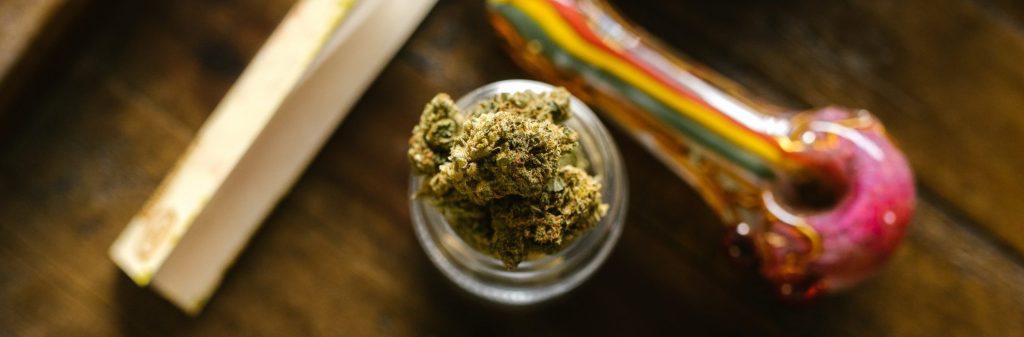 Top view of cannabis in a jar sitting next to a colorful pipe and rolling papers on wooden surface