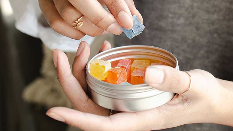 Hand holding round tin filled with red, yellow, orange, and blue cubed edibles.