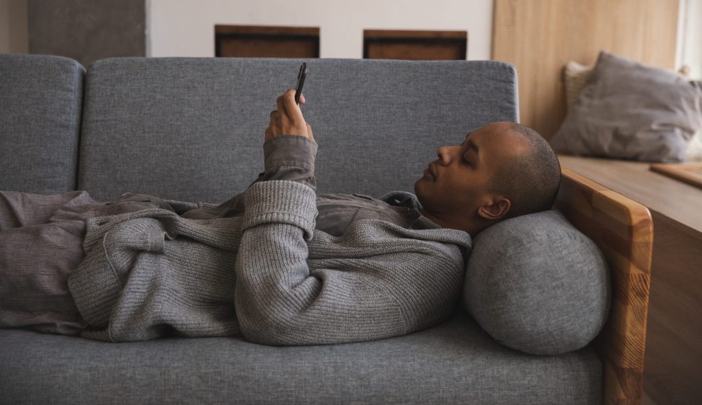 Person lying on gray couch, using cell phone, wearing gray sweater