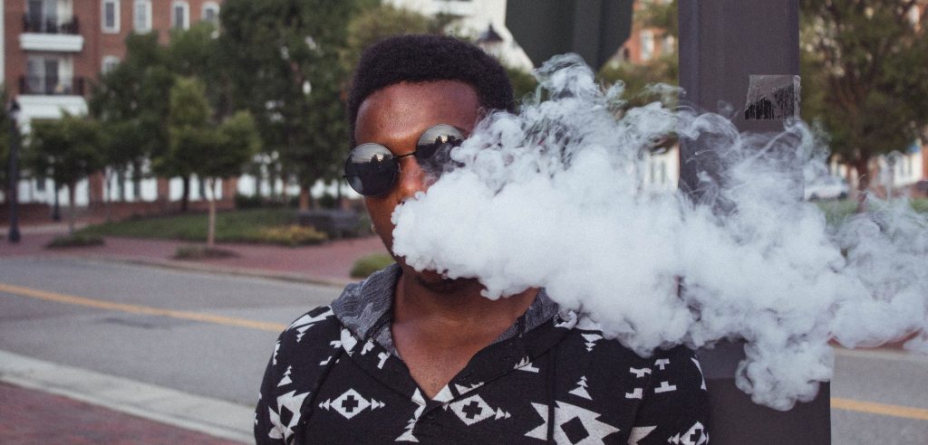 Person in black sunglasses and black and white patterned shirt, standing on a street corner, blowing a cloud of smoke